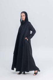 Woman wearing a black linen abaya with textured fabric detail and matching hood, styled for modest modern wear.
