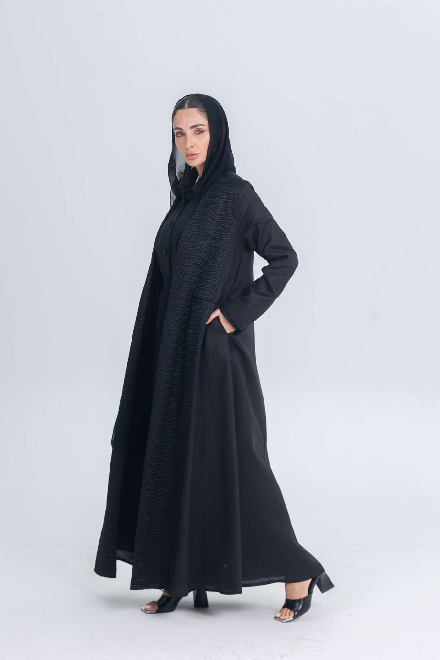 Woman wearing a black linen abaya with textured fabric detail and matching hood, styled for modest modern wear.
