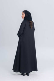 Woman wearing a black linen abaya with textured fabric detail and matching hijab, standing against a plain background.