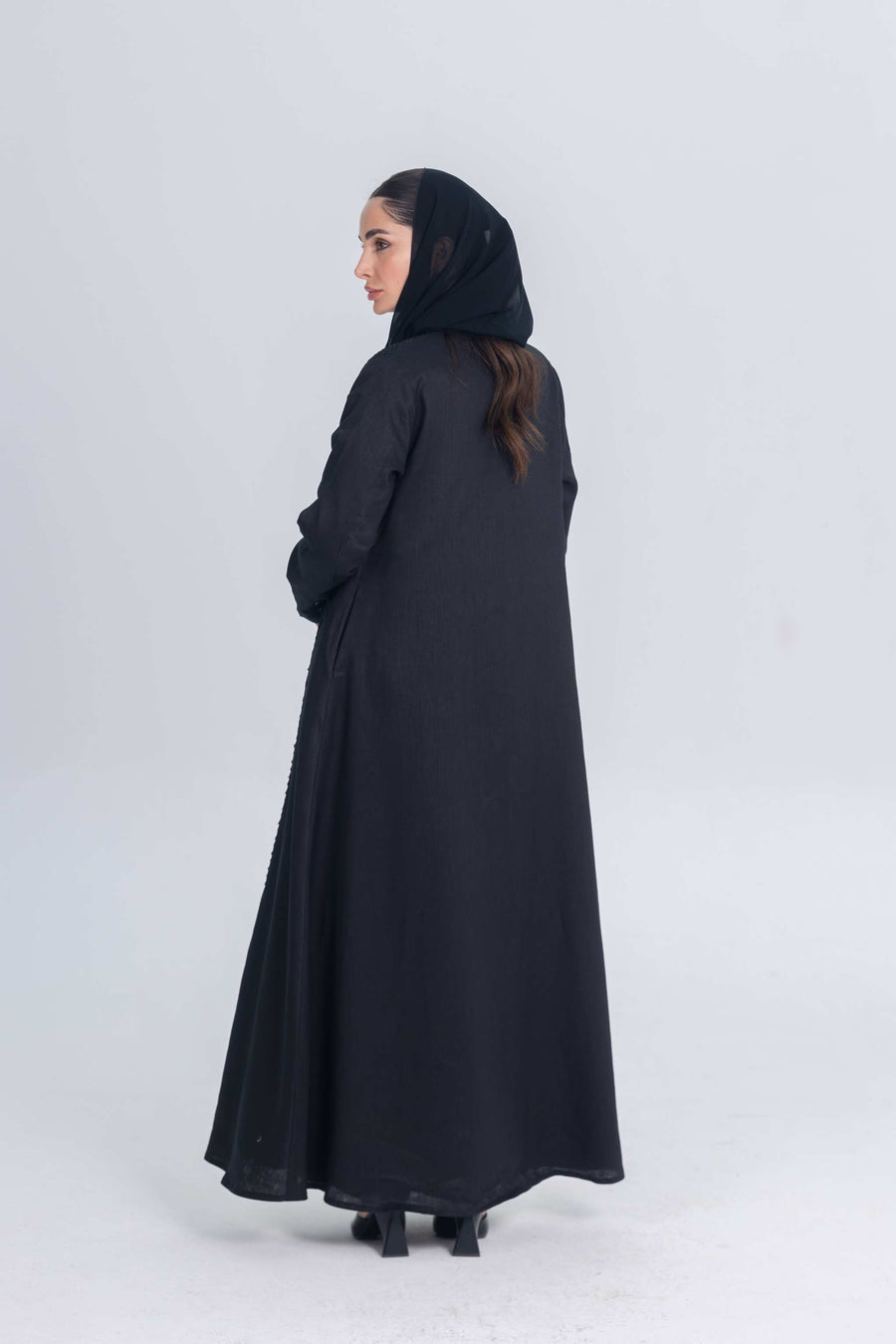 Woman wearing a black linen abaya with textured fabric detail and matching hijab, standing against a plain background.