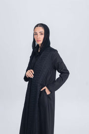Woman wearing a black linen abaya with textured fabric detail and matching inner set, styled with a black headscarf on a plain background