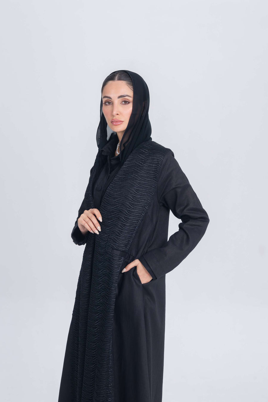 Woman wearing a black linen abaya with textured fabric detail and matching inner set, styled with a black headscarf on a plain background