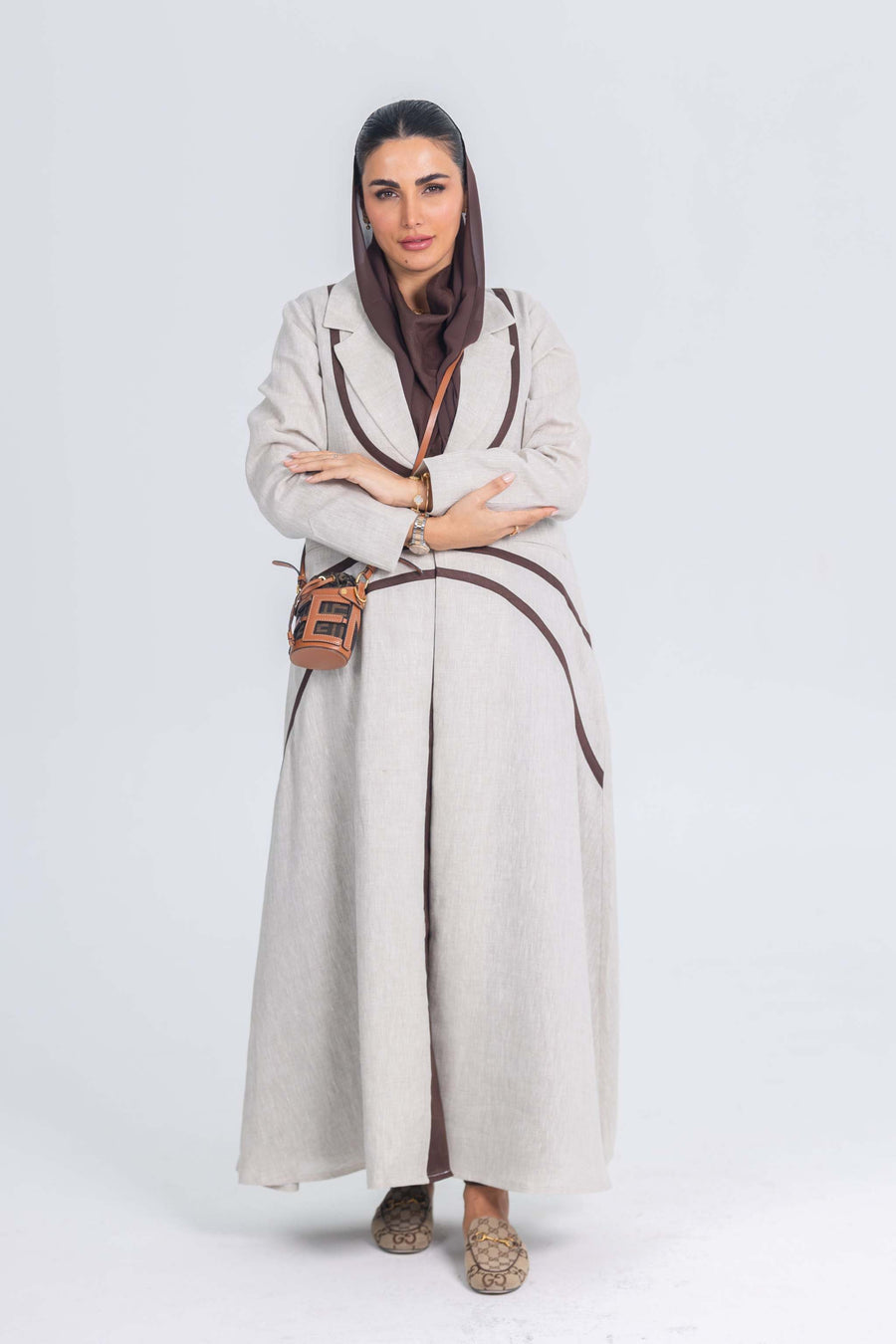 Woman wearing beige linen abaya with brown brim detailing holding a small brown handbag