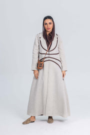 Woman wearing beige linen abaya with rich brown brim detailing and a crossbody bag against plain background
