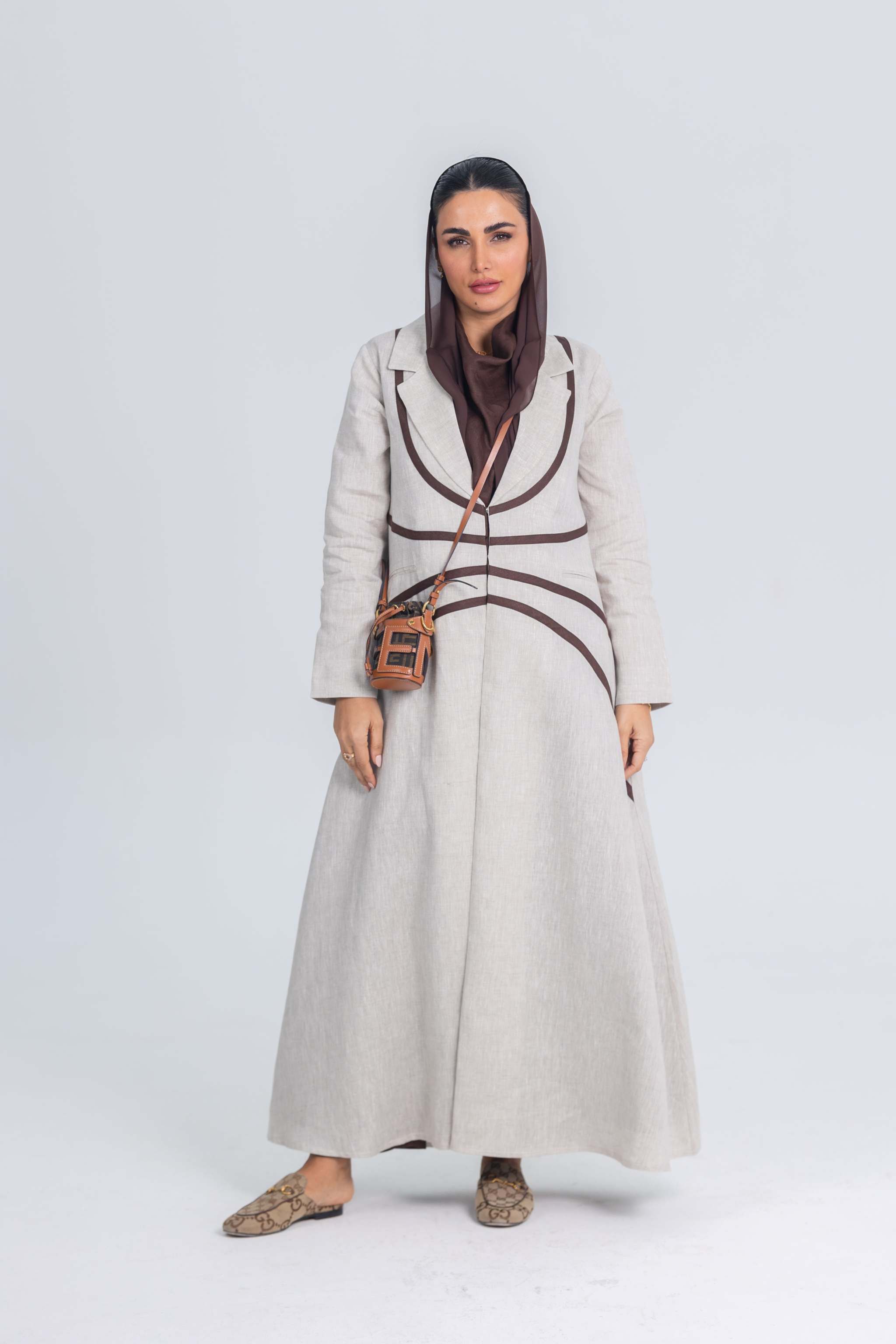 Woman wearing beige linen abaya with rich brown brim detailing and a crossbody bag against plain background