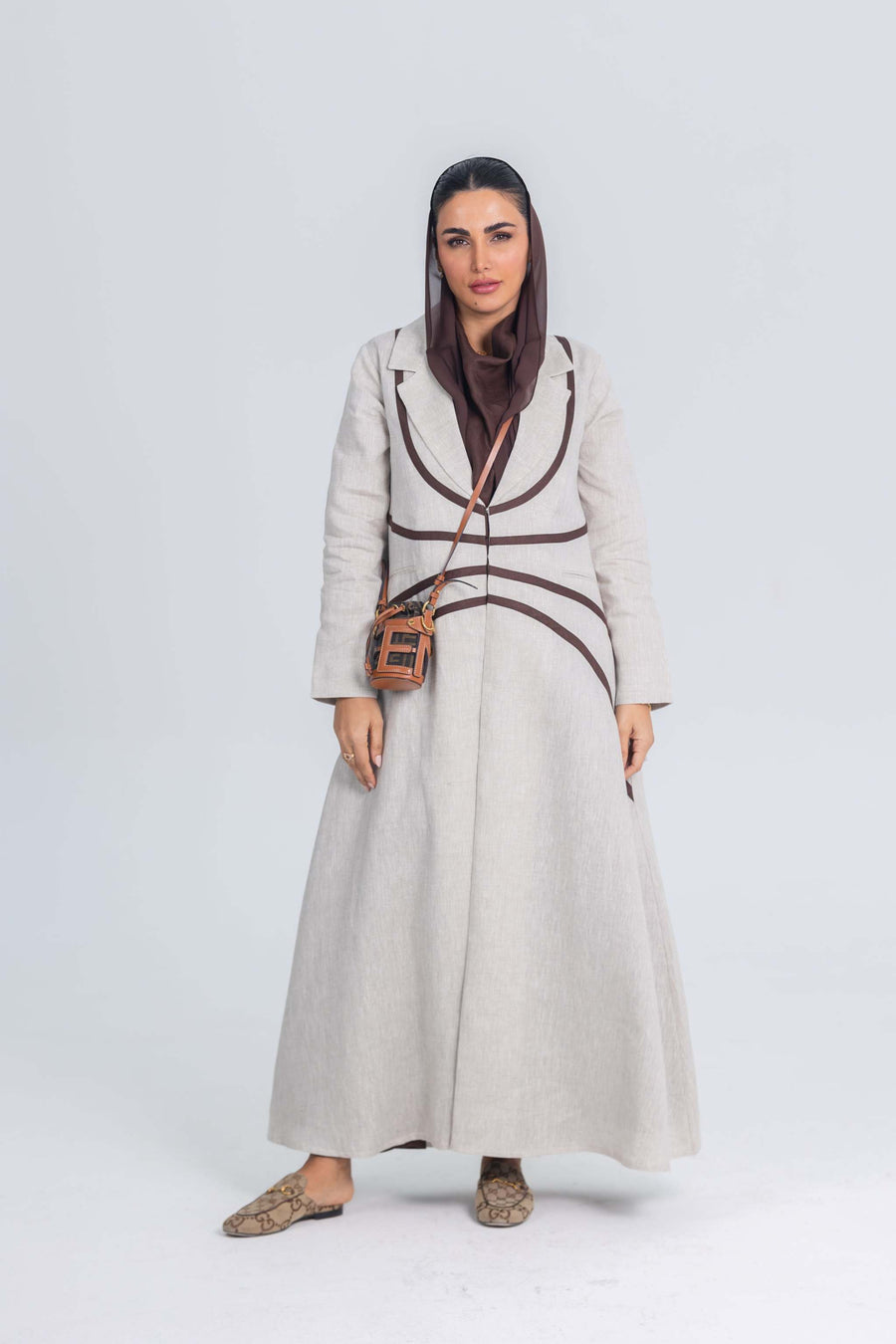 Woman wearing beige linen abaya with rich brown brim detailing and a crossbody bag against plain background