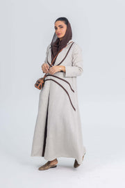 Beige linen abaya with brown brim detailing worn by woman, featuring elegant contrast lines and soft drape for refined daytime or office wear.