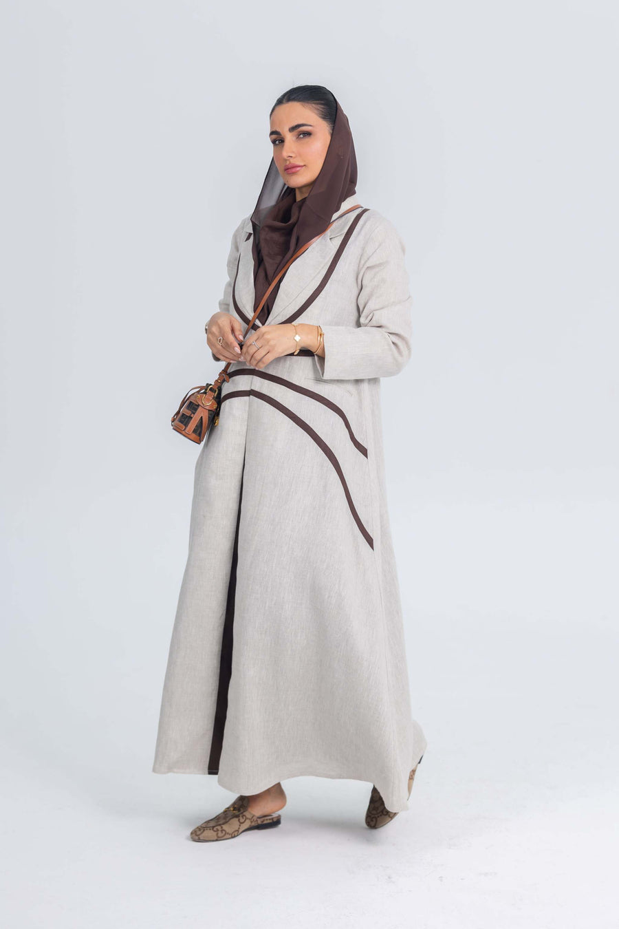 Beige linen abaya with brown brim detailing worn by woman, featuring elegant contrast lines and soft drape for refined daytime or office wear.