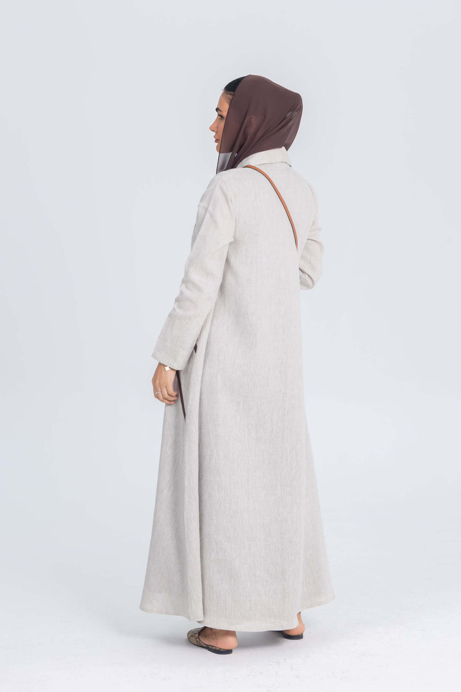 Beige linen abaya with brown brim worn by woman in brown hijab, standing side view on white background