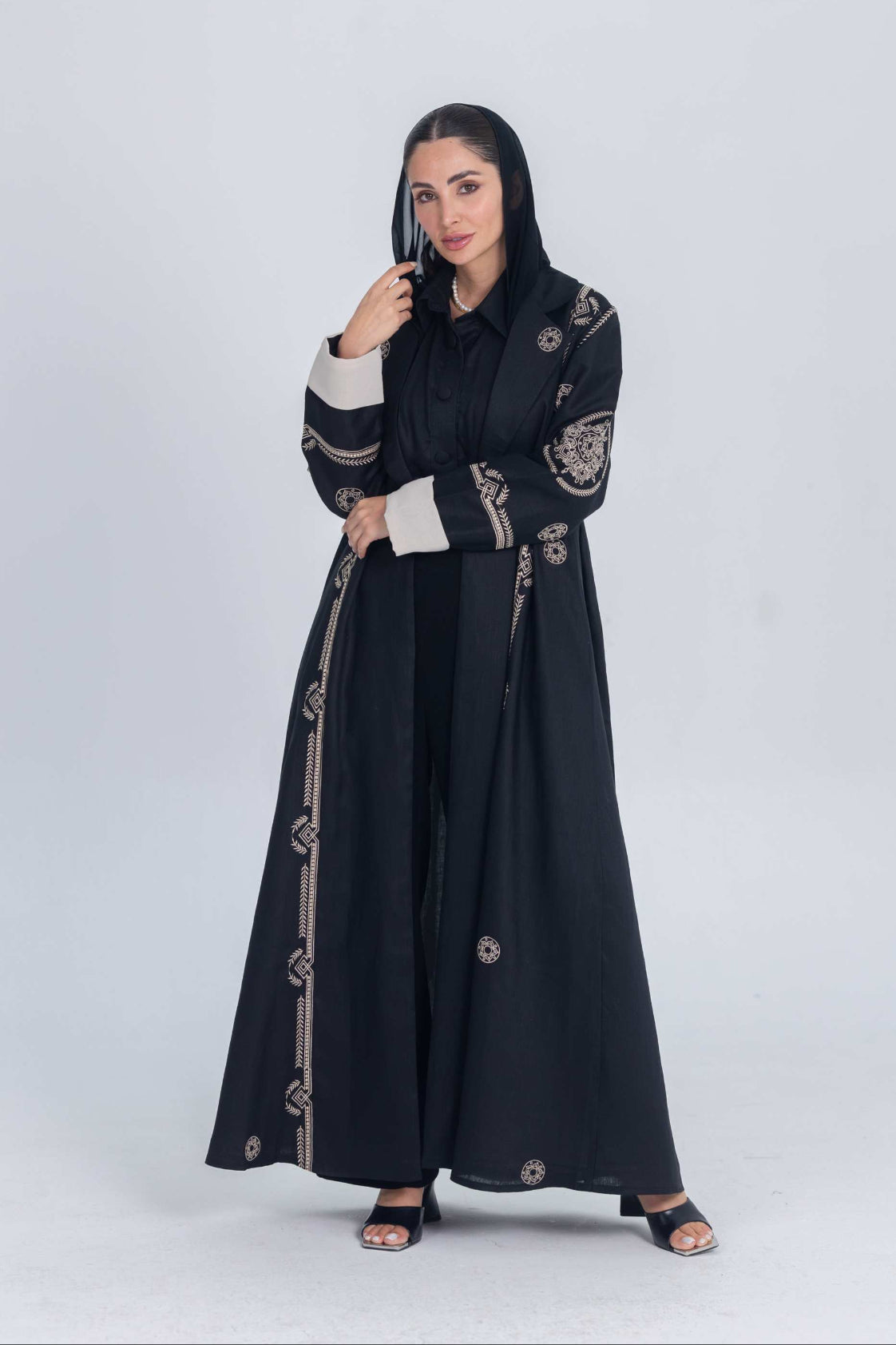Woman wearing a black luxury linen abaya with beige circular embroidery and cuff details standing against a plain background