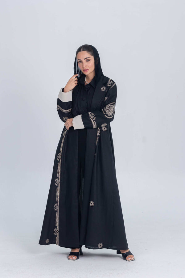 Woman wearing a black luxury linen abaya with beige circular embroidery and cuff details standing against a plain background