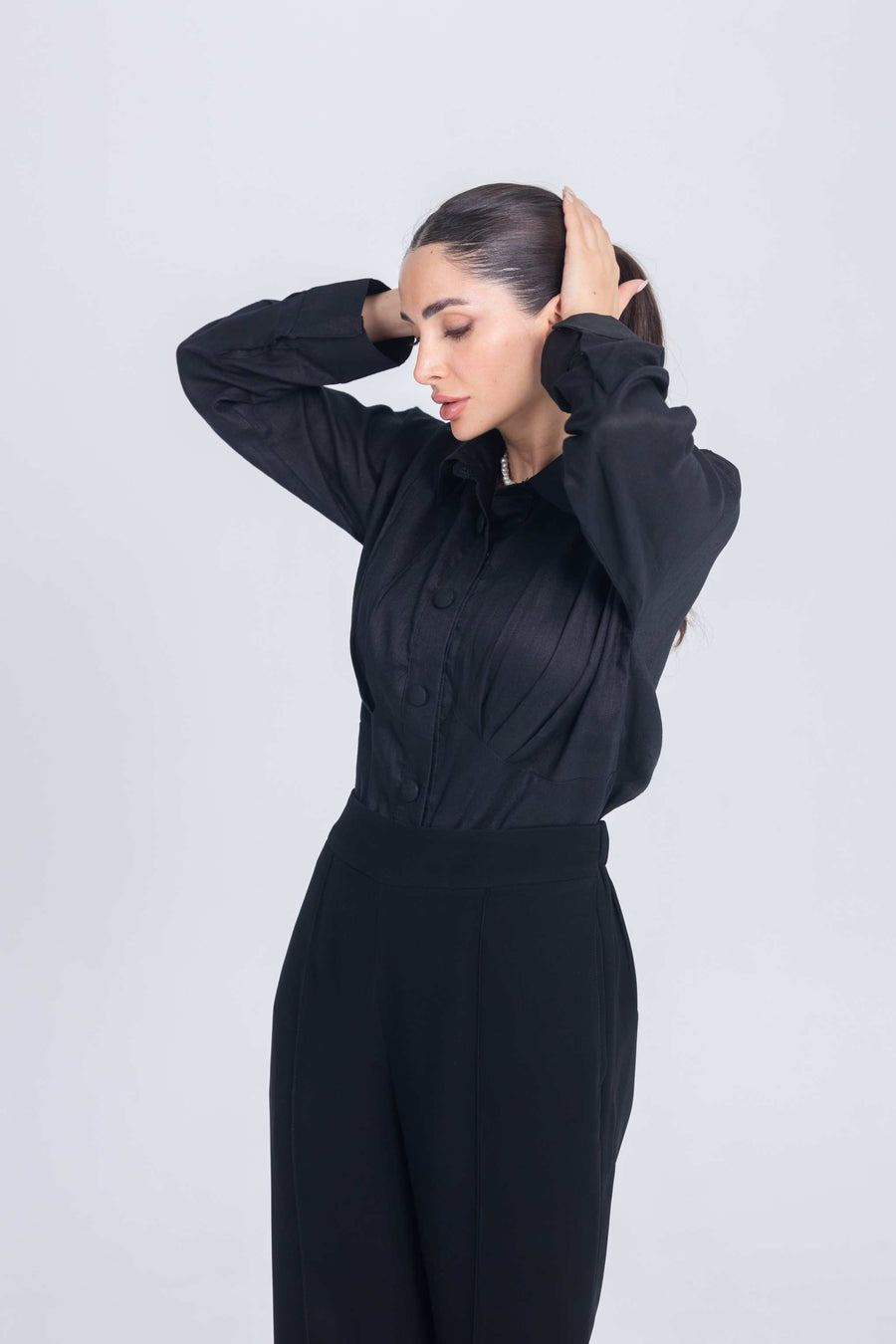 Woman wearing elegant black shirt with pleated chest detail and matching black pants against white background