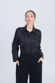 Woman wearing black shirt with chest pleats and black trousers standing against plain background
