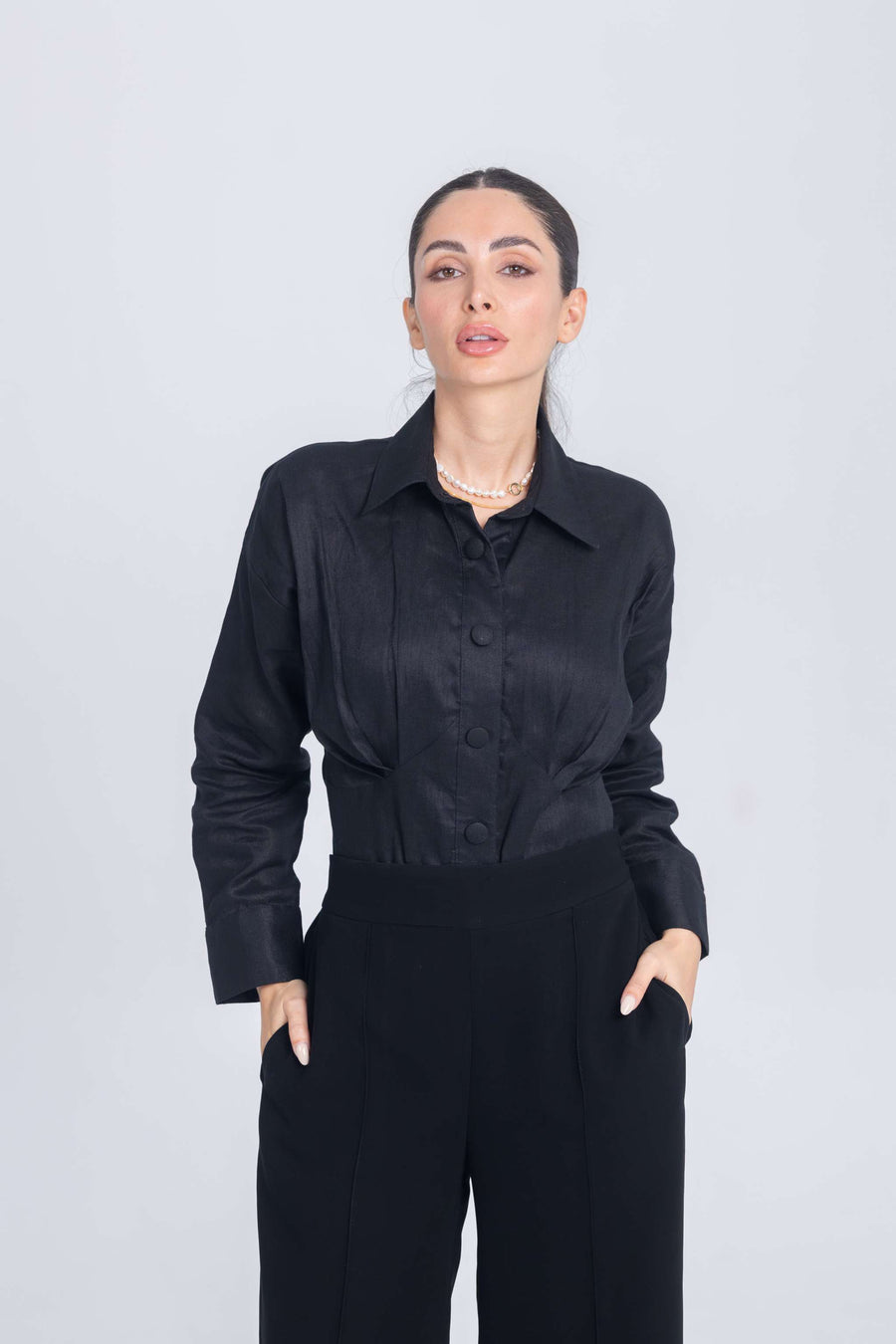 Woman wearing black shirt with chest pleats and black trousers standing against plain background