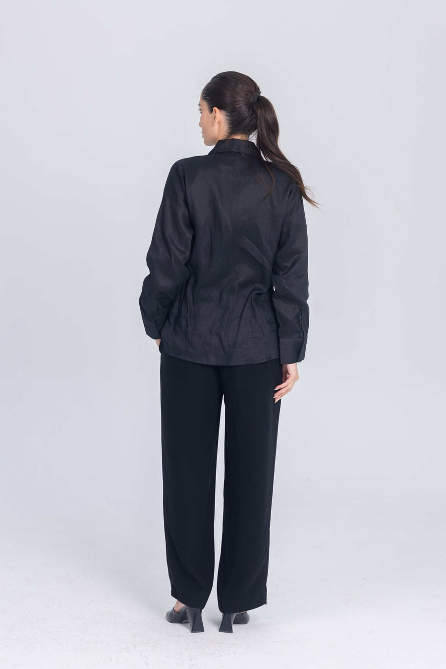 Woman wearing black Essence Folds shirt with pleated chest detail and black pants, back view
