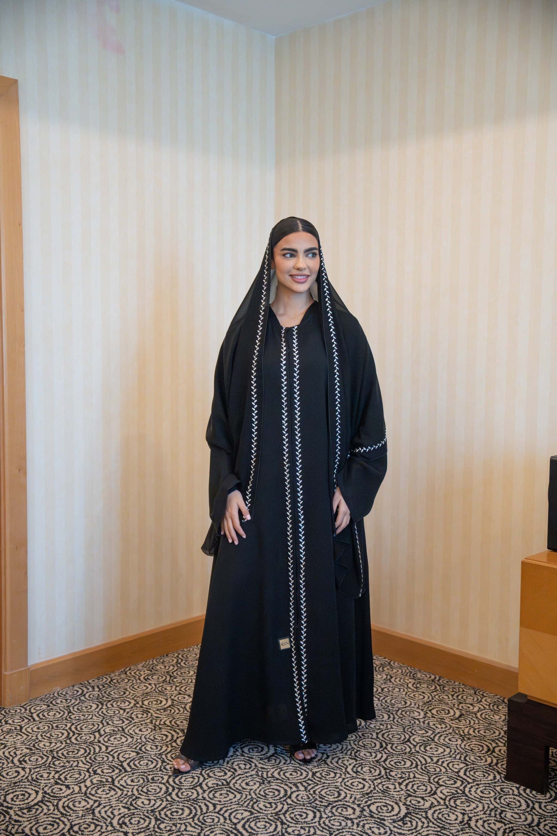 Woman wearing sleek black abaya with white embroidery and round neckline standing indoors.