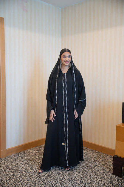 Woman wearing sleek black abaya with white embroidery and round neckline standing indoors.