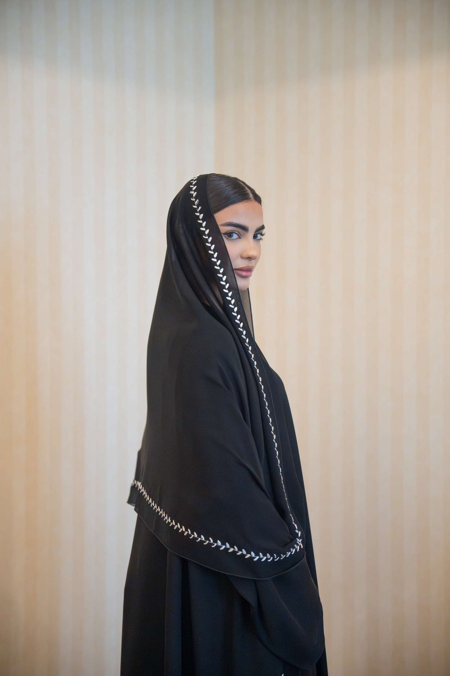 Woman wearing a sleek black abaya with delicate white embroidery along the edges and cuffs, paired with a matching embroidered sheila.