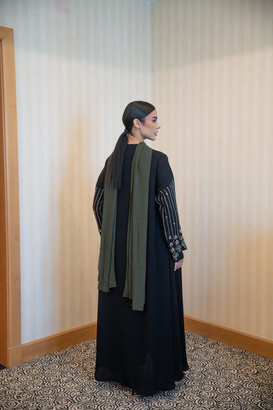 Back view of a woman wearing a Classic Straight-Cut Black Abaya with floral embroidered sleeves and olive green trim, featuring a loose fit and front buttons.
