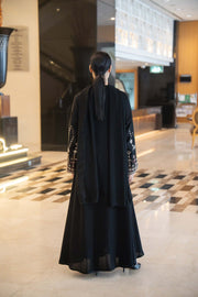 Back view of a flowing black abaya with straight cut, round neckline trimmed in soft brown, and wide flared sleeves embroidered in coffee brown and off-white.