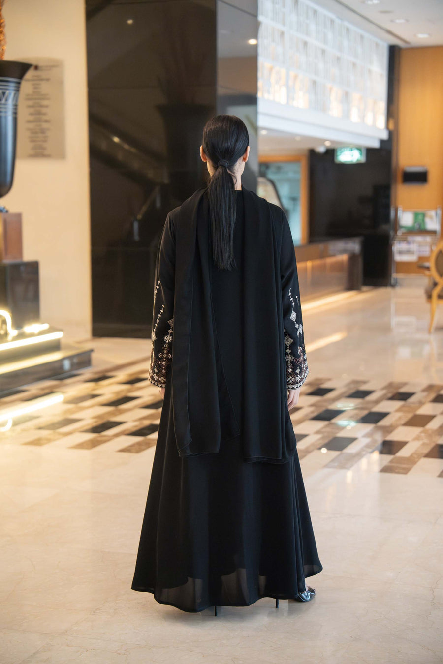 Back view of a flowing black abaya with straight cut, round neckline trimmed in soft brown, and wide flared sleeves embroidered in coffee brown and off-white.