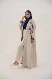 Woman wearing a soft beige premium linen abaya with navy floral embroidery and gold accents, featuring navy inner cuffs and a loose open bisht cut.