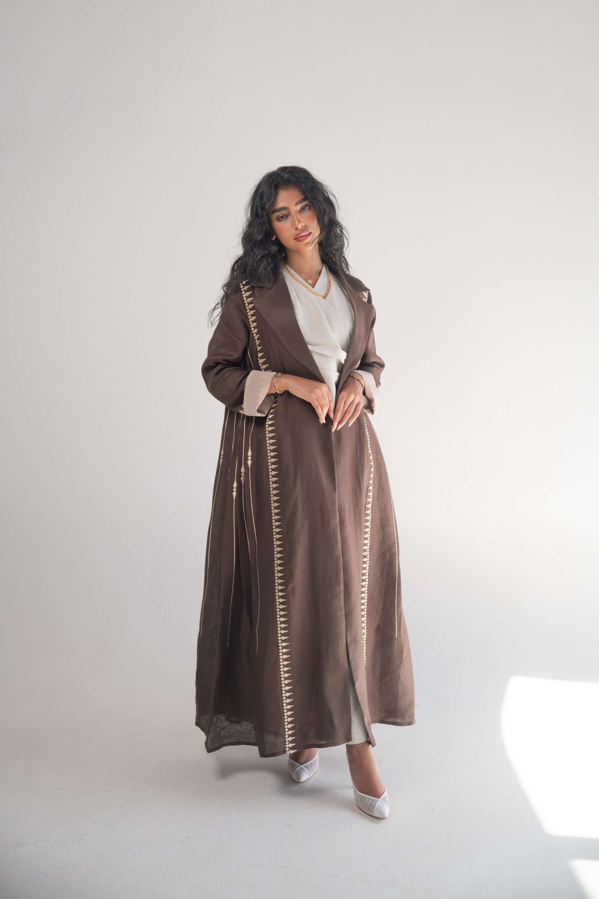 Woman wearing a dark brown linen blazer abaya with beige triangular embroidery and blazer collar, styled with a light beige chiffon inner layer and subtle back pleats.