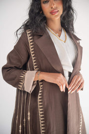 Woman wearing a dark brown linen blazer abaya with beige triangular embroidery and blazer collar, styled with a light beige chiffon inner layer and subtle back pleats.