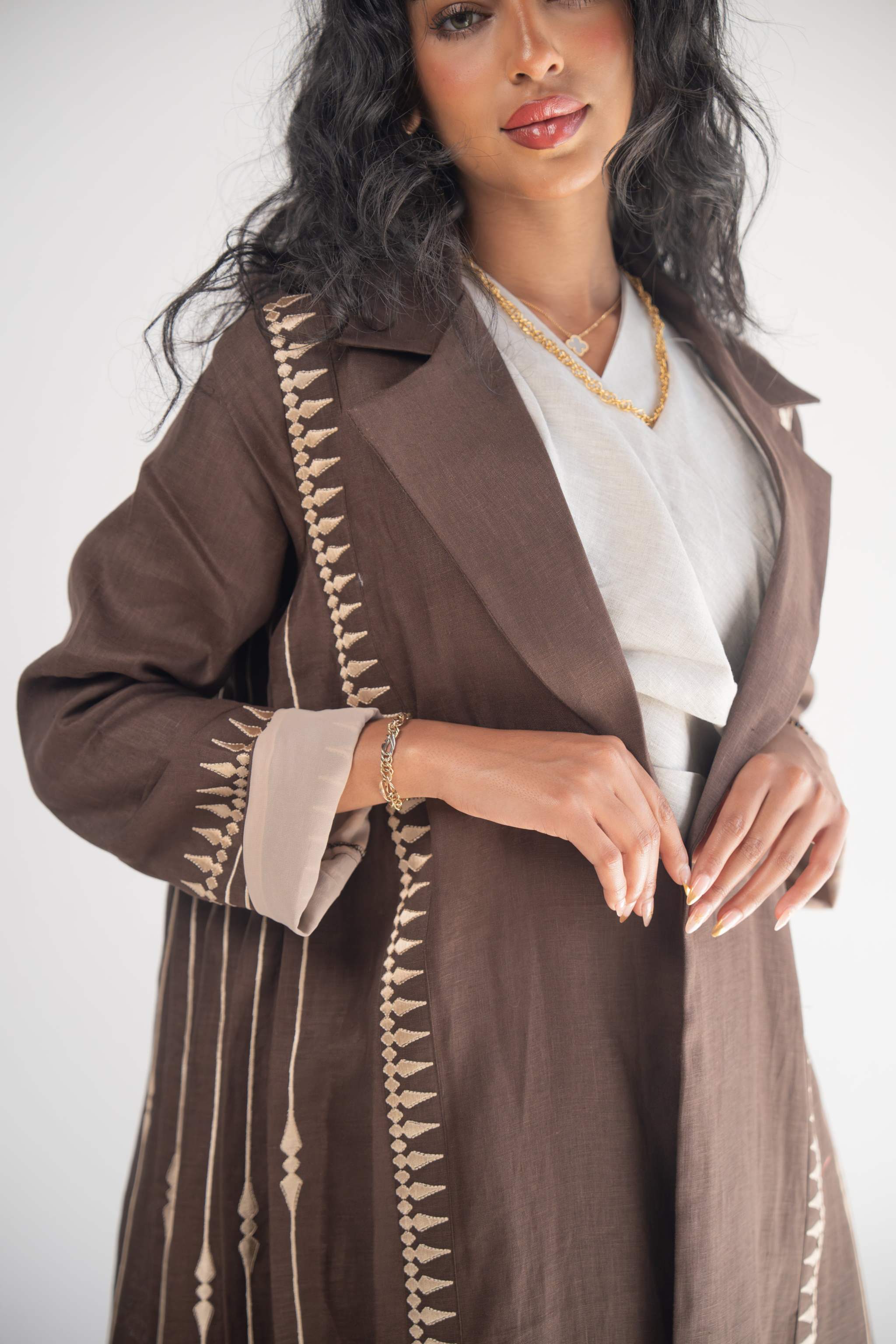 Woman wearing a dark brown linen blazer abaya with beige triangular embroidery and blazer collar, styled with a light beige chiffon inner layer and subtle back pleats.