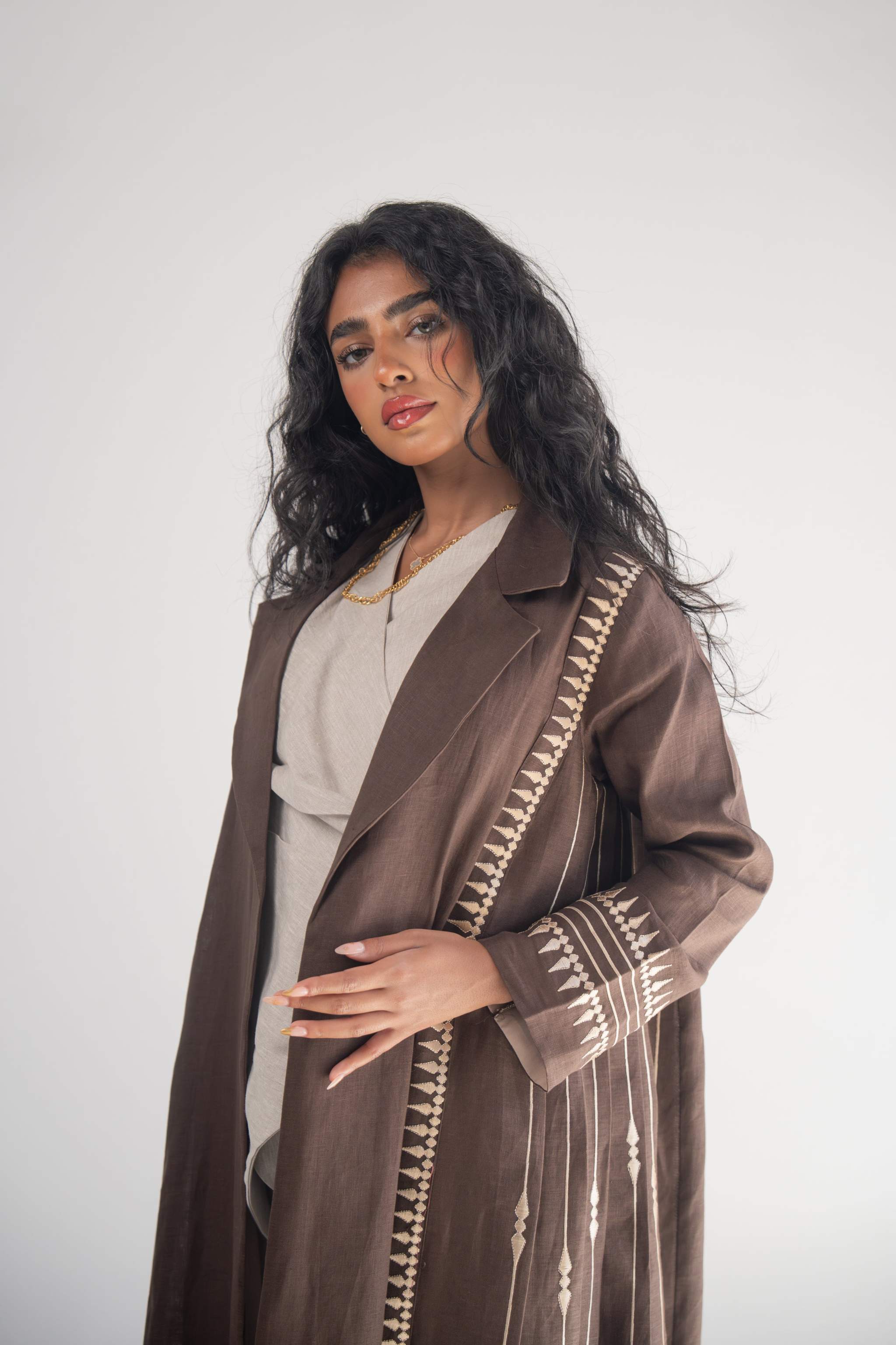 Woman wearing a dark brown linen blazer abaya with beige triangular embroidery and blazer collar, styled with a light beige chiffon inner layer and subtle back pleats.