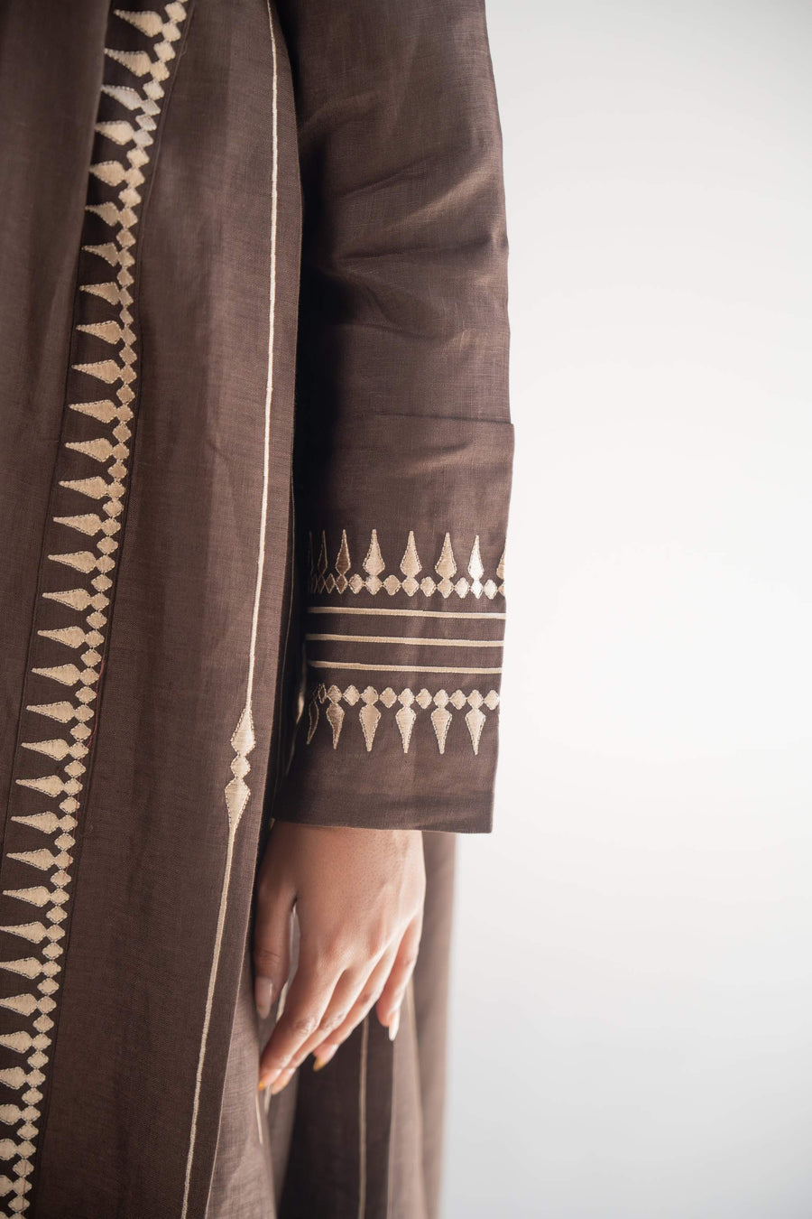Woman wearing a dark brown linen blazer abaya with beige triangular embroidery and blazer collar, styled with a light beige chiffon inner layer and subtle back pleats.