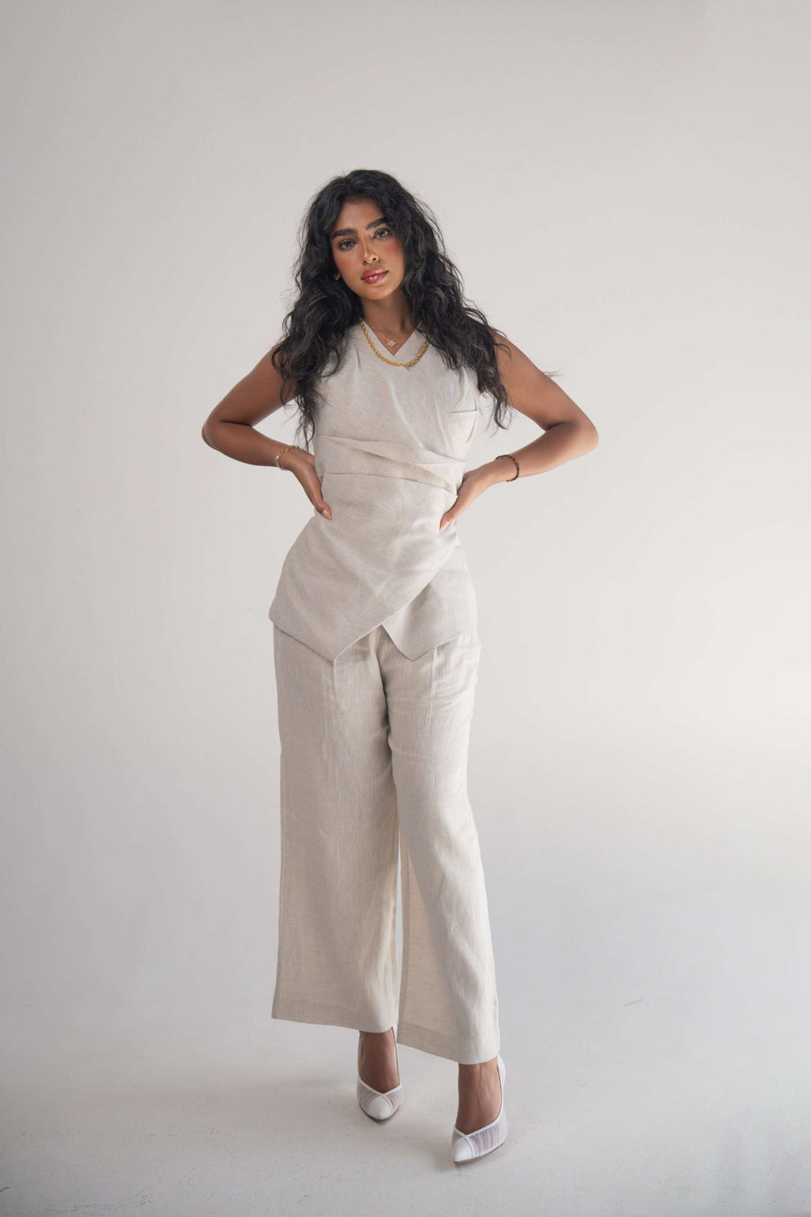 Woman wearing sleeveless Almond Beige Blouse with front wrap and soft pleats, paired with matching wide-leg pants and white heels.