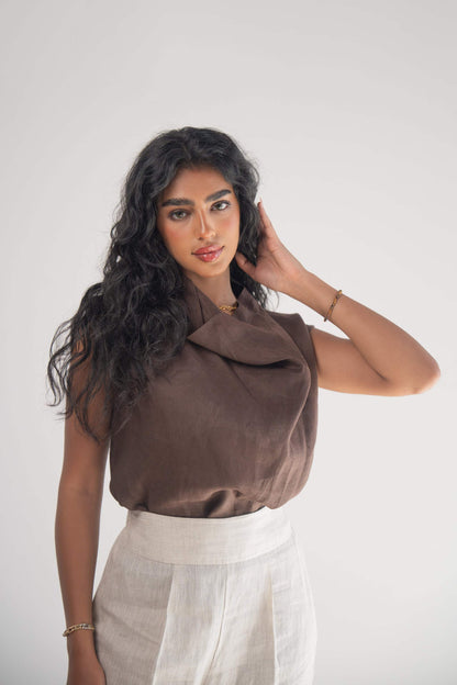 Woman wearing a sleeveless cocoa linen blouse with a draped neckline and white linen pants against a plain background