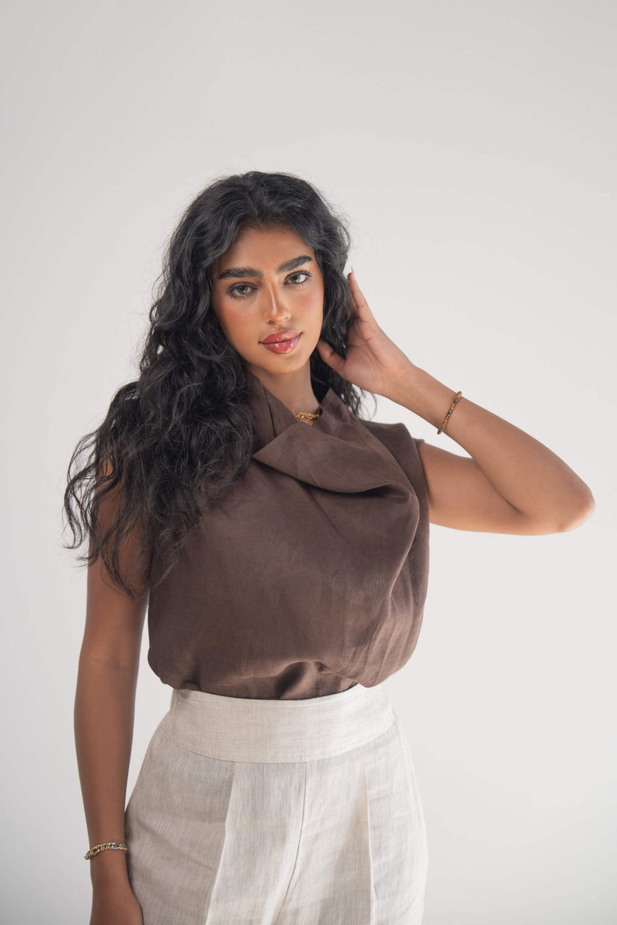 Woman wearing a sleeveless cocoa linen blouse with a draped neckline and white linen pants against a plain background