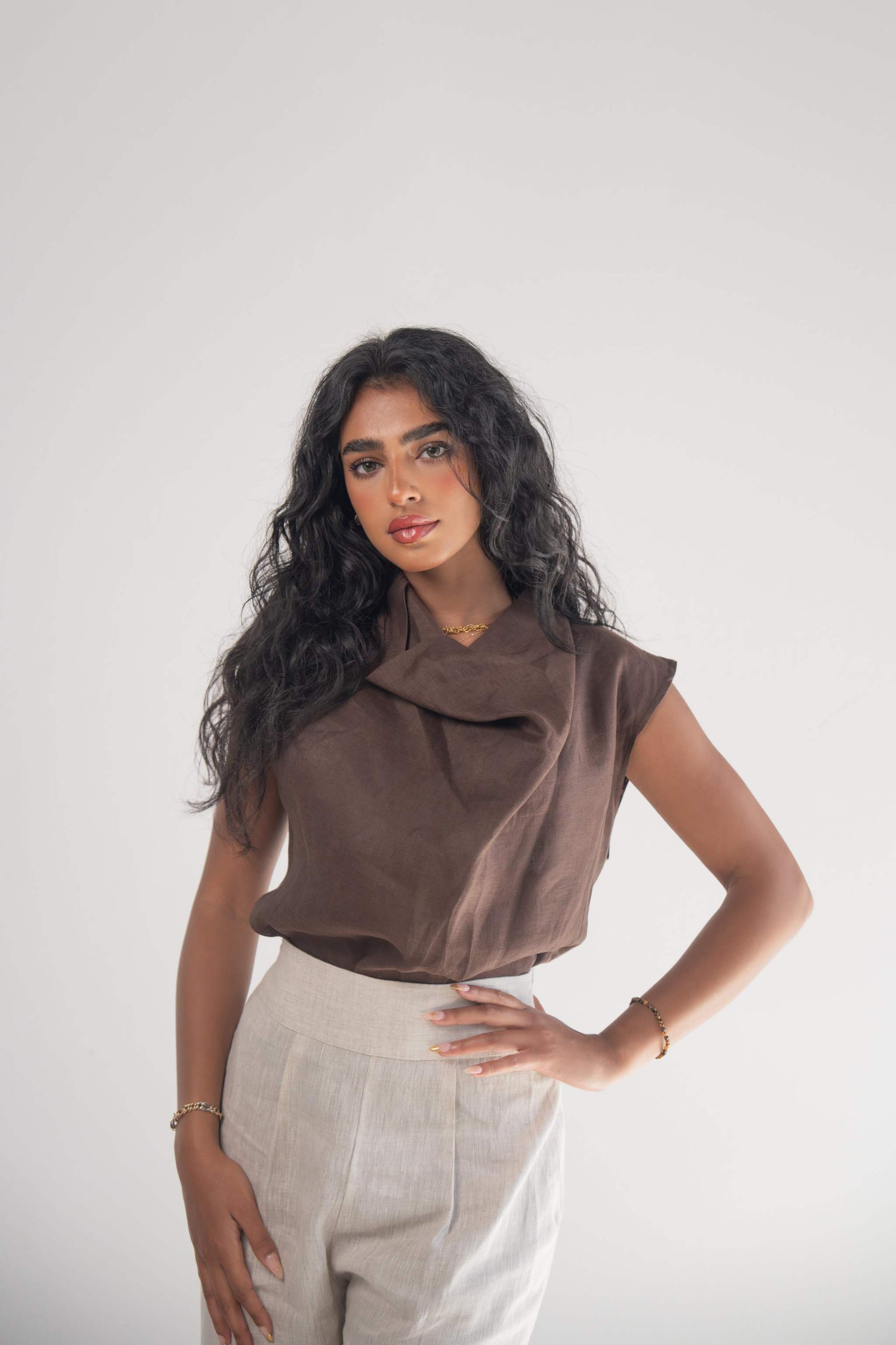 Woman wearing sleeveless cocoa brown linen blouse with draped neckline and subtle pleats paired with light-colored pants