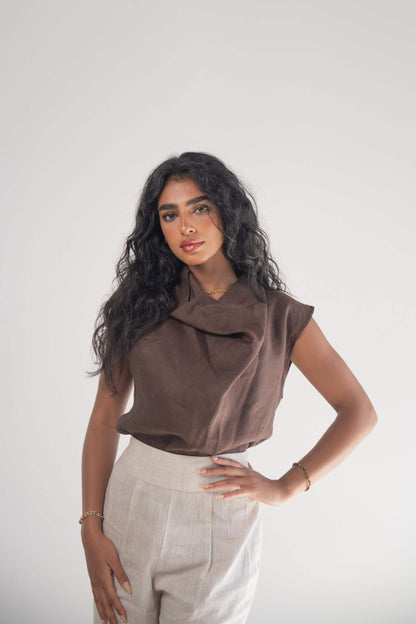Woman wearing sleeveless cocoa brown linen blouse with draped neckline and subtle pleats paired with light-colored pants