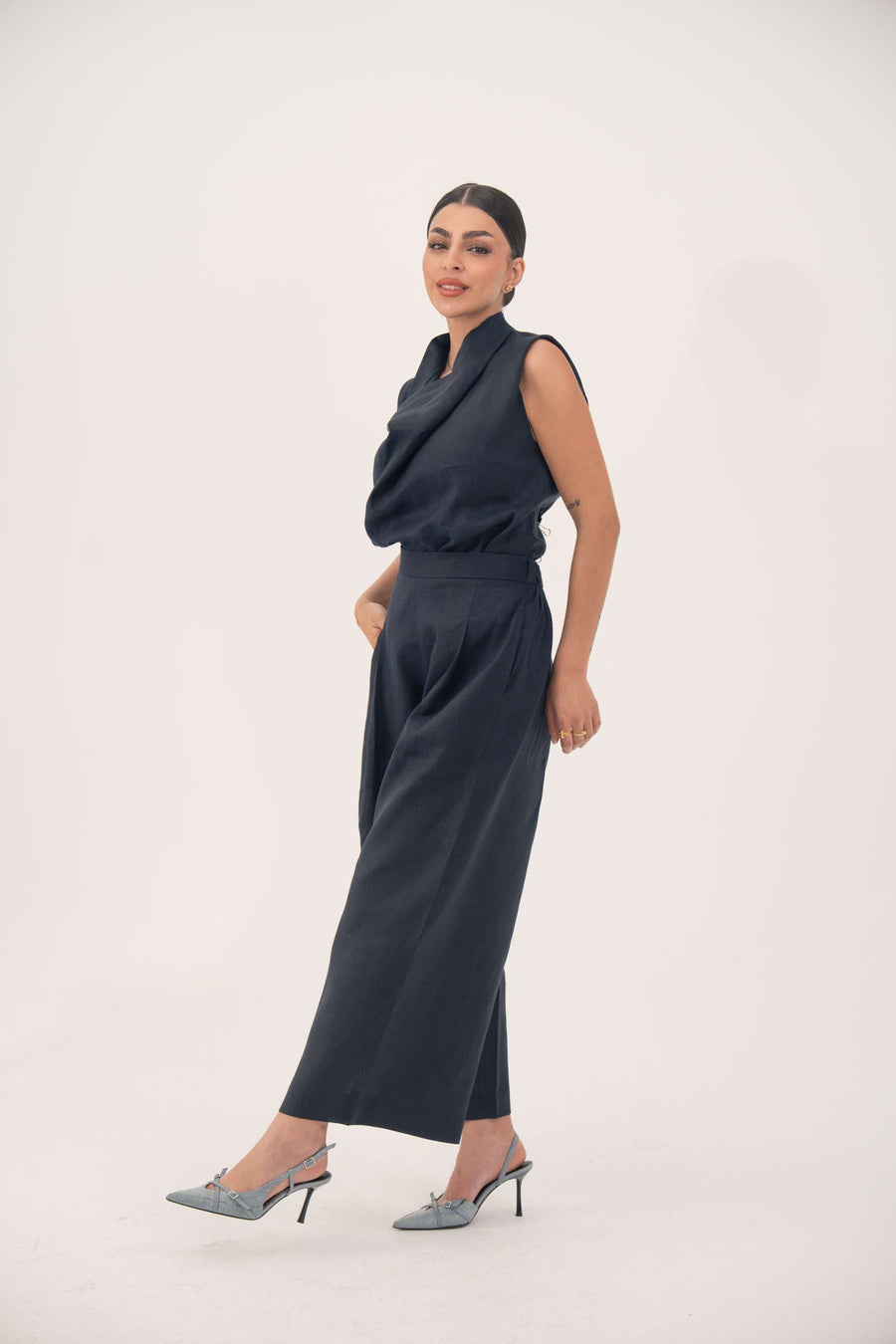 Woman wearing sleeveless navy blue linen blend blouse with draped cowl neckline and loose fit tucked into high-waisted pants
