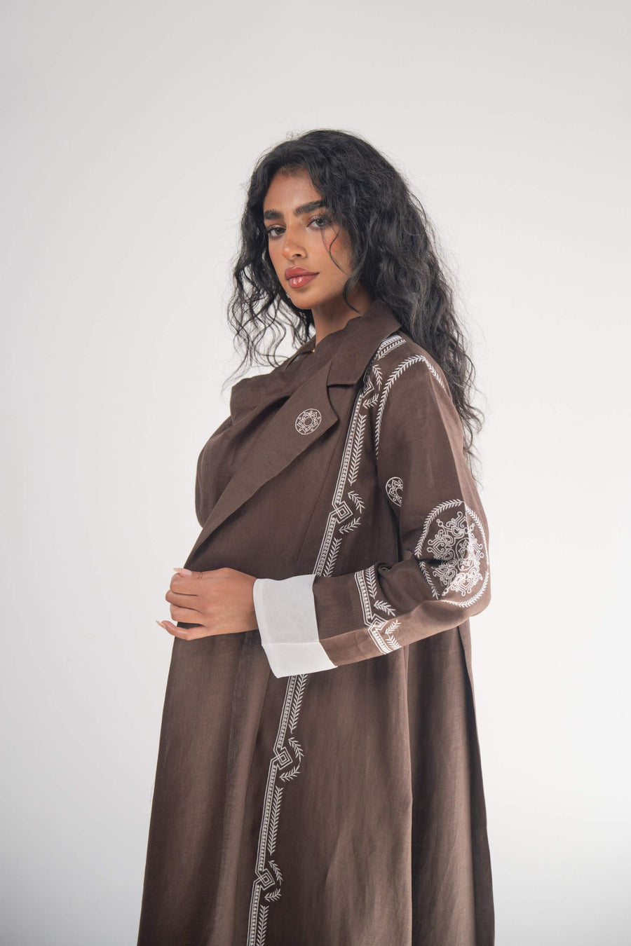Woman wearing a cocoa brown linen blazer abaya with white geometric and circular embroidery on sleeves and sides, featuring a classic blazer lapel and white chiffon lining.