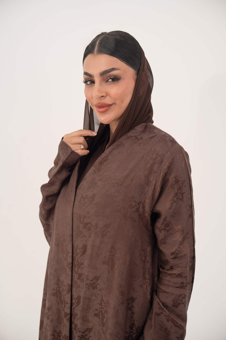 Woman wearing a warm cocoa brown jacquard silk abaya with subtle floral pattern and long sleeves, featuring a relaxed A-line cut and clean edges.