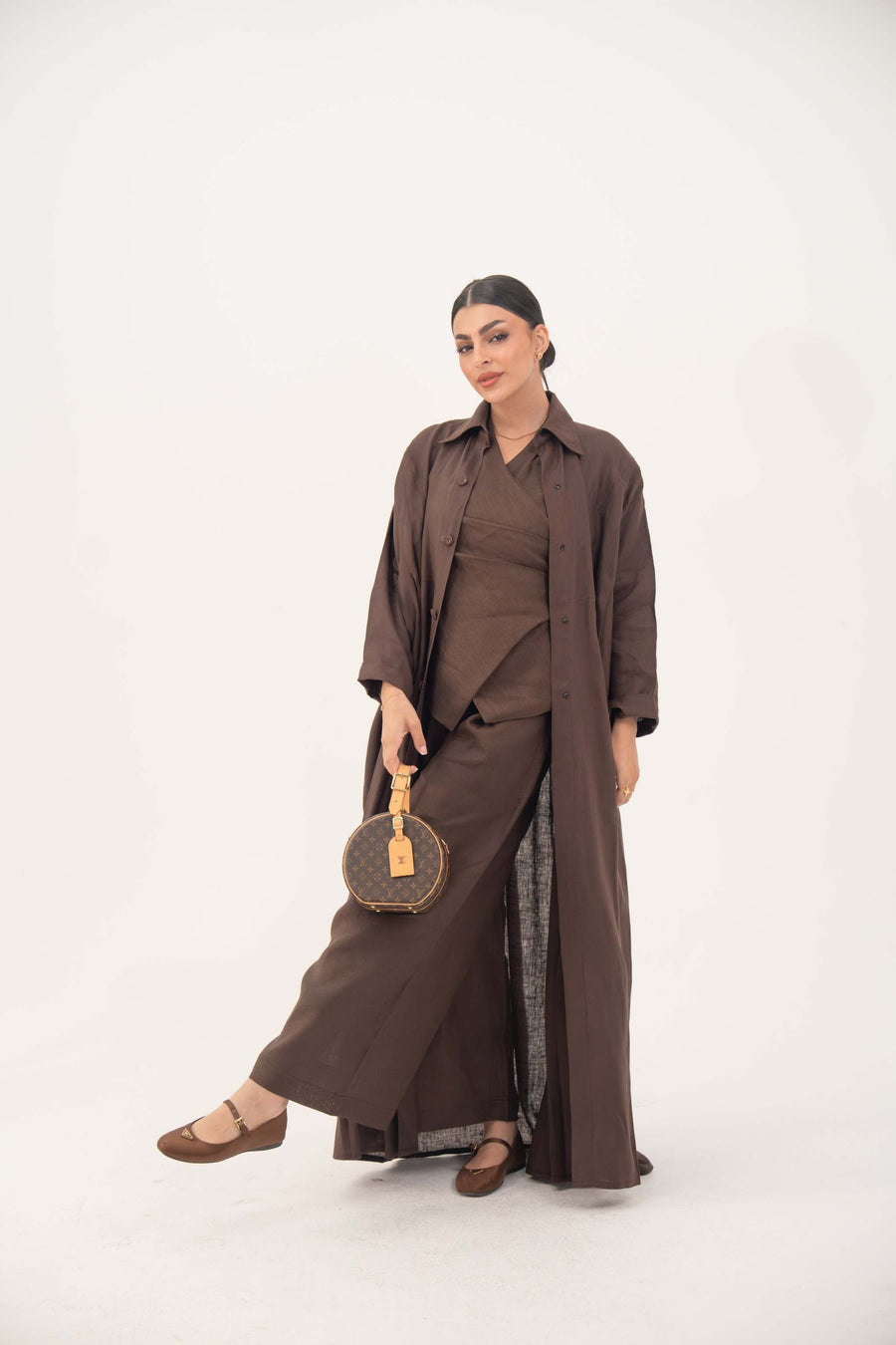 Woman wearing deep cocoa brown linen shirt-inspired abaya with classic collar and tonal buttons, holding round handbag.