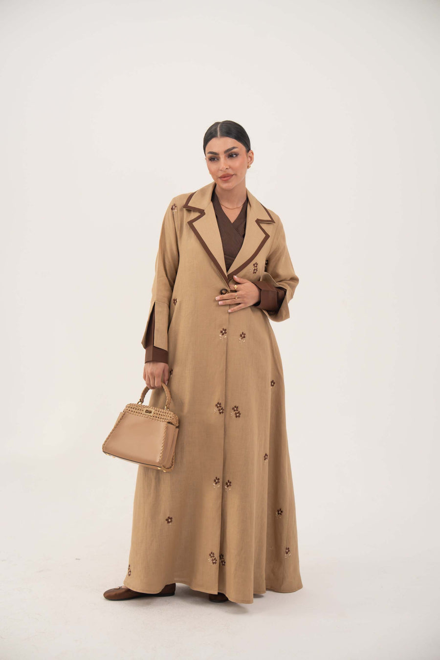 Dark brown A-line linen abaya with beige pleated panels and cream floral embroidery, featuring draped lapel and full lining for comfort.