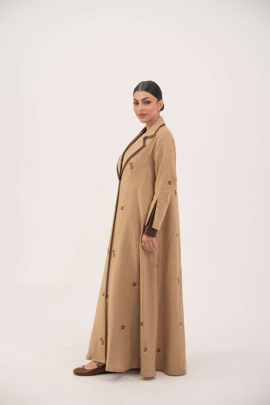 Dark brown linen abaya with beige pleated side panels and cream floral embroidery, featuring a draped lapel and A-line silhouette.