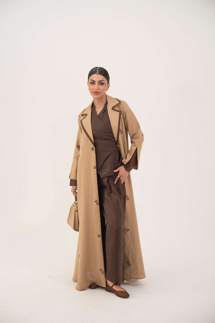 Woman wearing a dark brown linen abaya with beige pleated side panels and cream floral embroidery, featuring a draped lapel and A-line silhouette