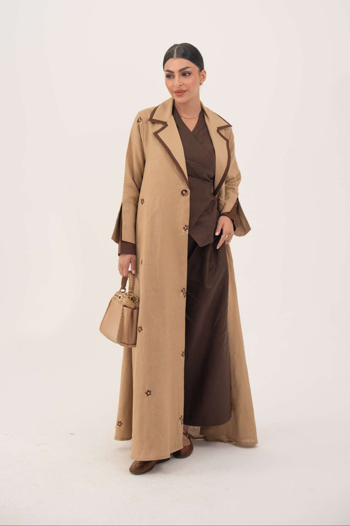 woman wearing cinnamon lightweight linen abaya with pleated side panels and floral embroidery carrying beige handbag