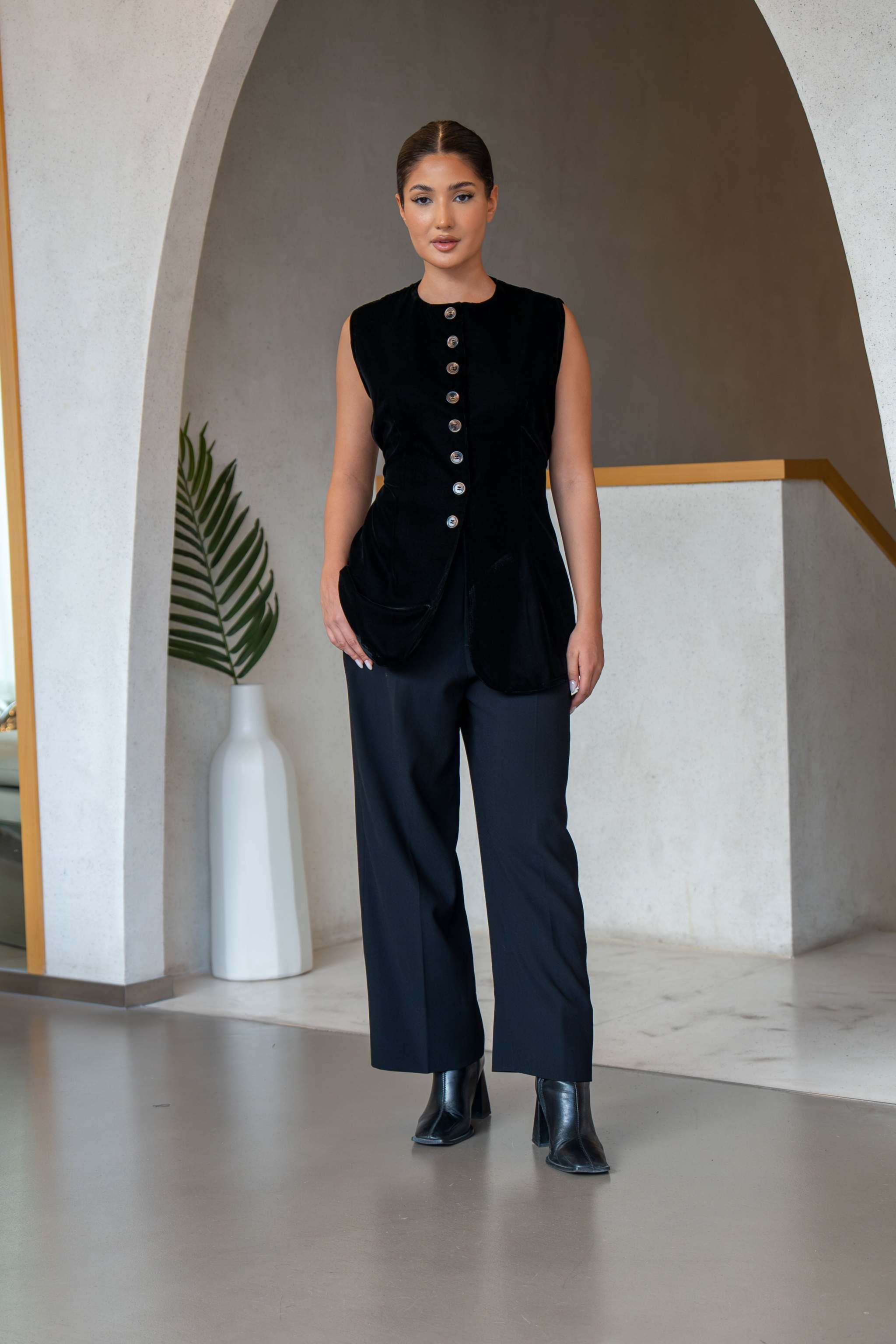 Woman wearing a sleek black velvet vest with front buttons styled with black pants and boots in a modern indoor setting