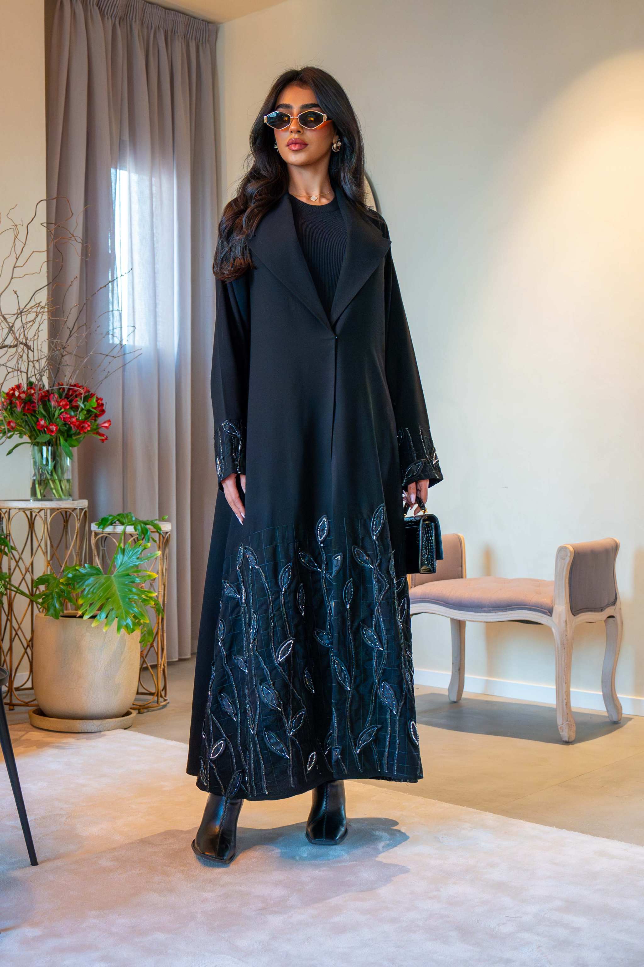 Woman wearing elegant black crepe Night Leaf abaya with leaf embroidery on cuffs and hem, paired with sunglasses and black boots in modern indoor setting