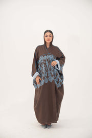 Woman wearing a loose mocha brown bisht abaya with blue shak embroidery on back and sleeves, light blue piping, and rolled cuffs.