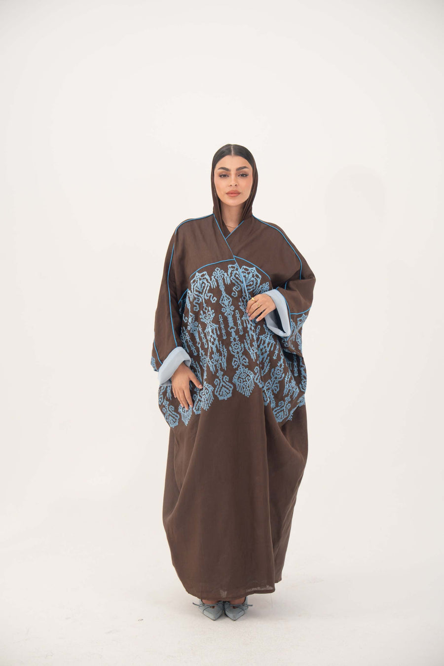 Woman wearing a loose mocha brown bisht abaya with blue shak embroidery on back and sleeves, light blue piping, and rolled cuffs.
