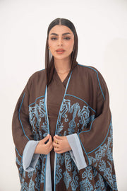 Woman wearing a mocha brown linen bisht abaya with blue shak embroidery, light blue piping, and rolled cuffs.
