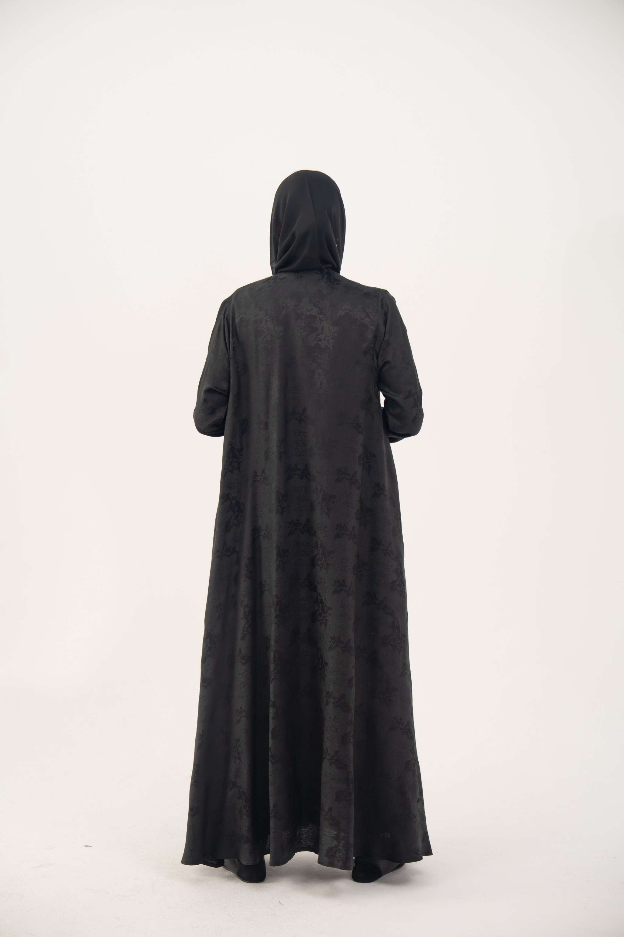 Back view of a black jacquard silk abaya with tone-on-tone floral weave, featuring a fluid A-line cut, relaxed shoulders, open front, straight sleeves, and discreet side pockets.